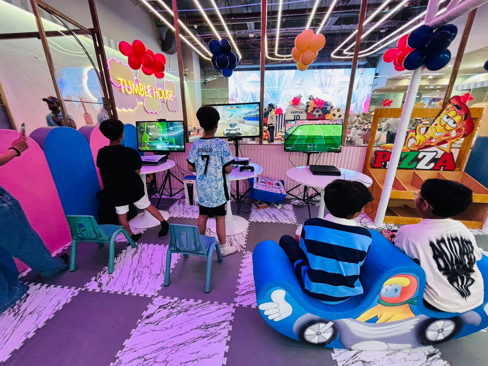 Children enjoying a multiplayer gaming experience at an event entertainment zone, playing racing and football video games on multiple screens in a colorful, themed indoor venue decorated with balloons and interactive seating.