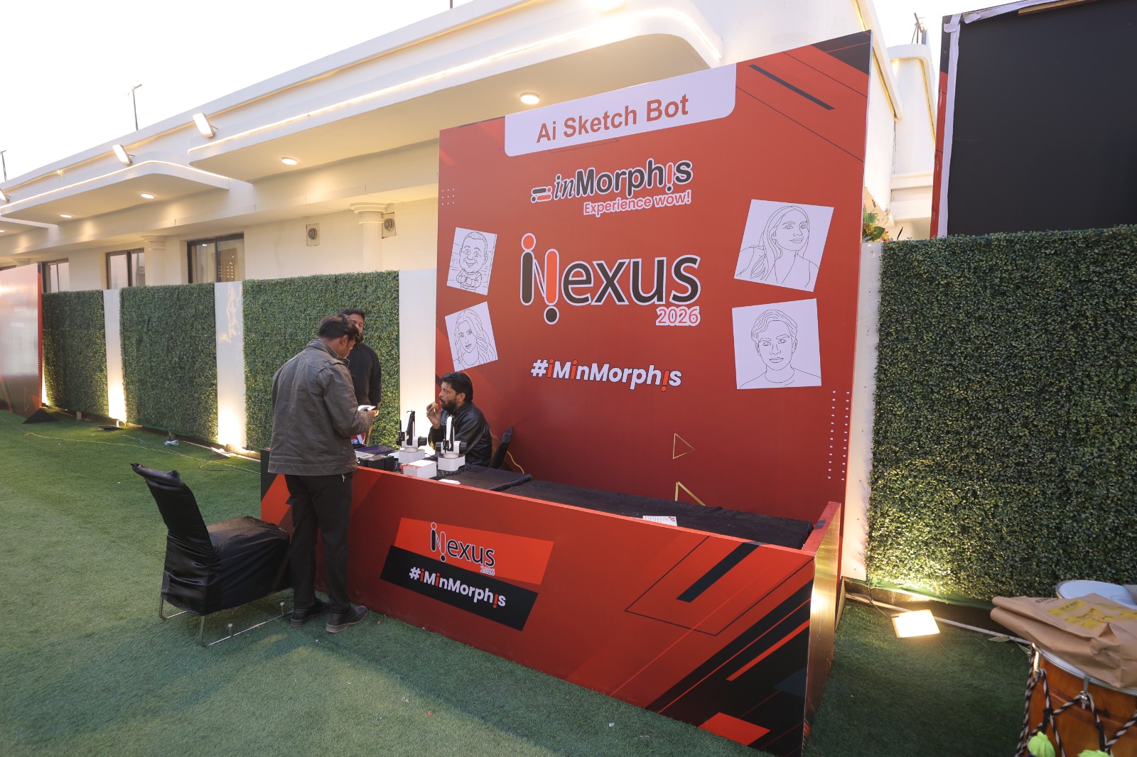 An AI Sketch Bot activation booth at iNexus 2026, featuring live AI-generated sketch portraits. Two team members operate laptops and sketching equipment in front of a branded red backdrop displaying sample portrait sketches and event branding.