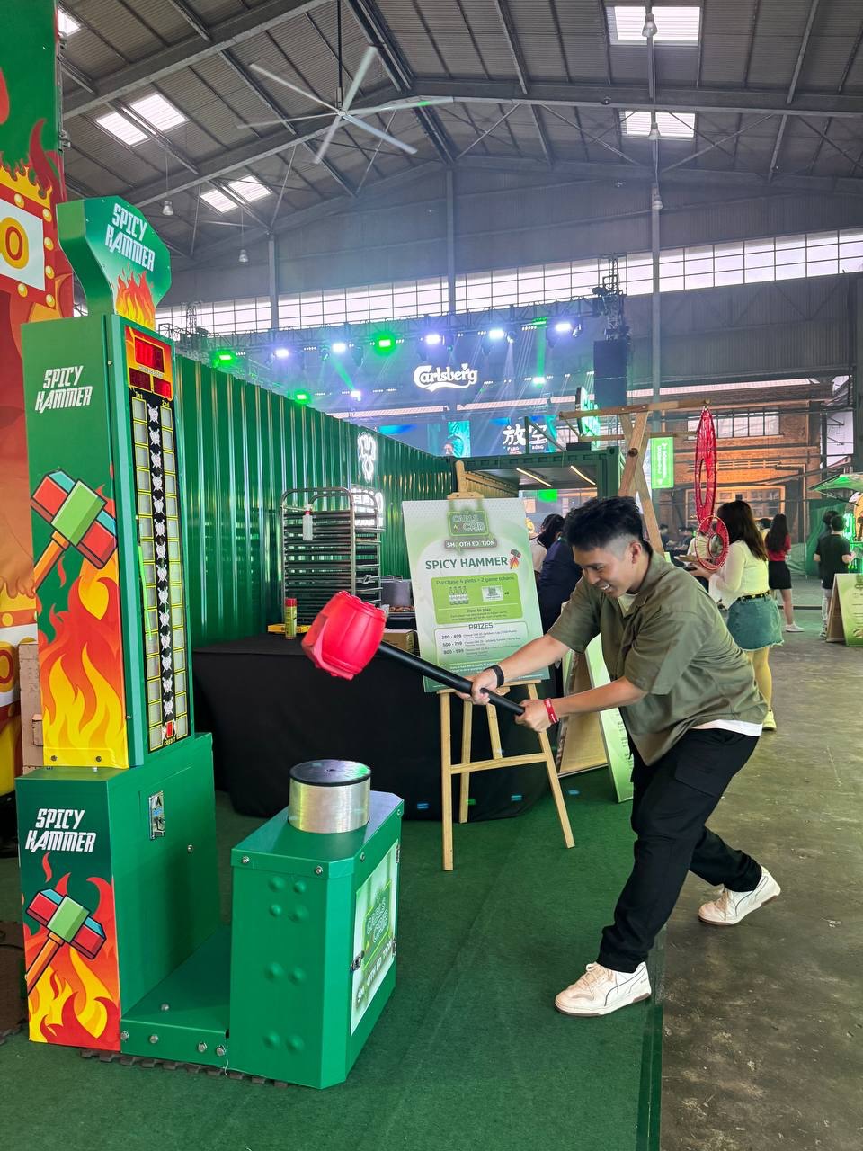 A participant playing the Spicy Hammer High Striker strength game at a branded event activation. The carnival-style hammer game is provided by Fabx Events for corporate events, brand activations, exhibitions, and roadshows.